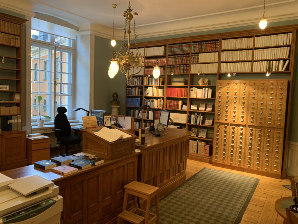 Rum med informationsdisk i mitten, grön matta som täcker ett trägolv. Ett kartotek till höger i bild, tillsammans med bokhyllor täcker en hel vägg. Ett fönster släpper in ljus och en ljuskrona i taket ger ett varmt gult sken.