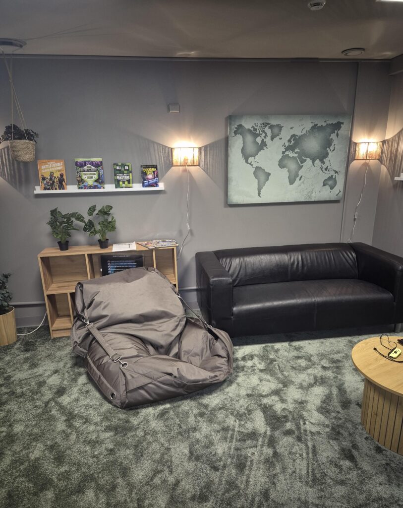 Ett rum med en ljusgrön heltäcknignsmatta, svart skinnsoffa, beige sittpuff, bokhylla, växter, mysiga vägglampor och en tavla med världskartan.