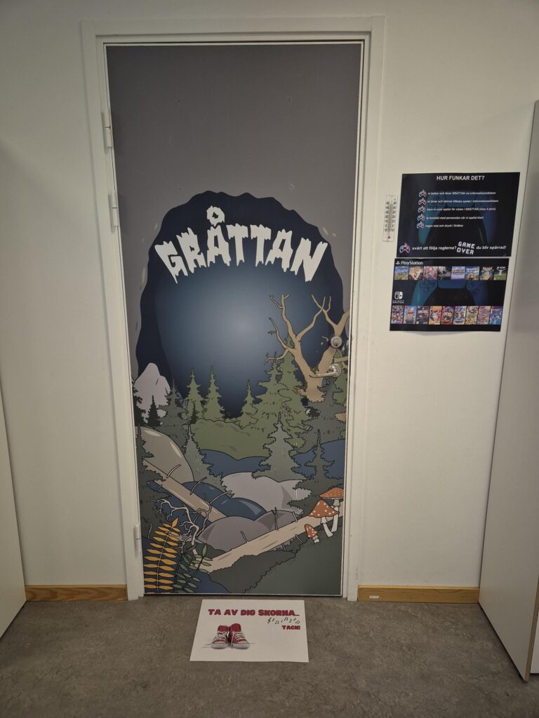 Dörren in till Gråttan är grå med en bild på en skogsmiljö och en grotta insprängd i det grå. På golvet en skoskylt om att ta av sig skorna.