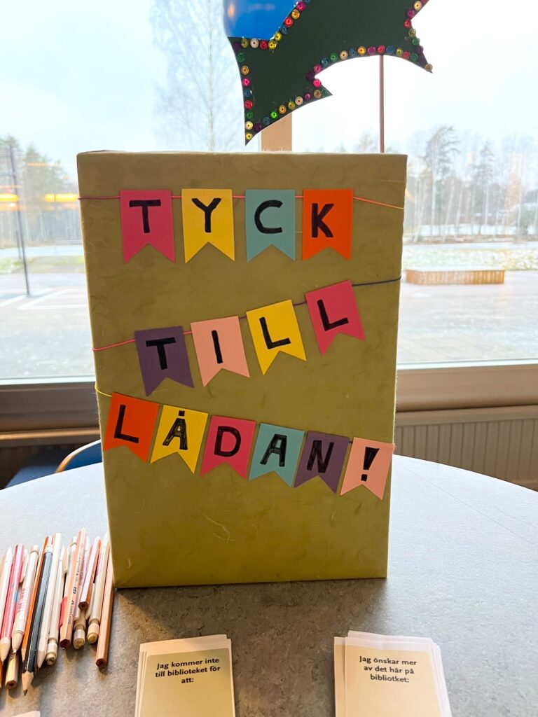 En låda står på ett bord och har texten TYCK TILL -LÅDAN i olika färger. Bredvid ligger en hög med pennor och några frågekort.