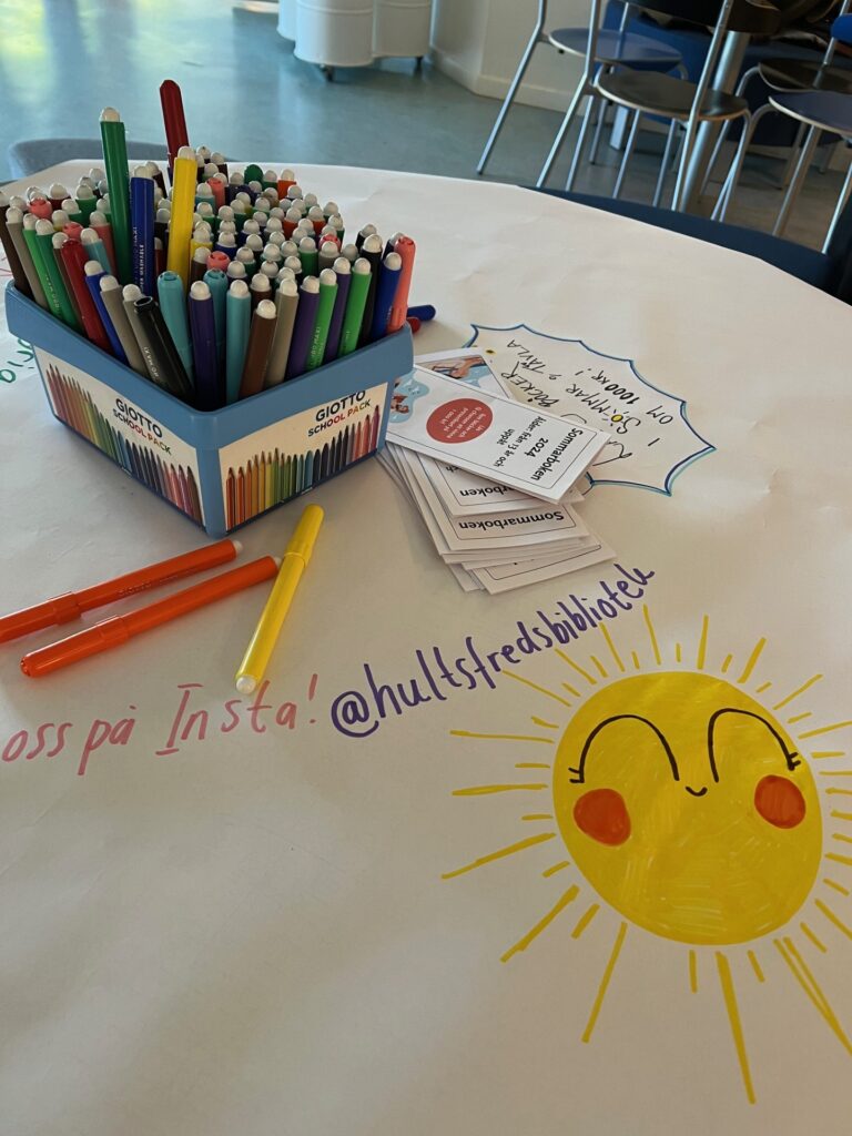 Ett bord med vit duk, där någon målat en glad sol samt instagramadressen @hultsfredbibliotek. På boret står en burk med tuschpennor i olika färger. 