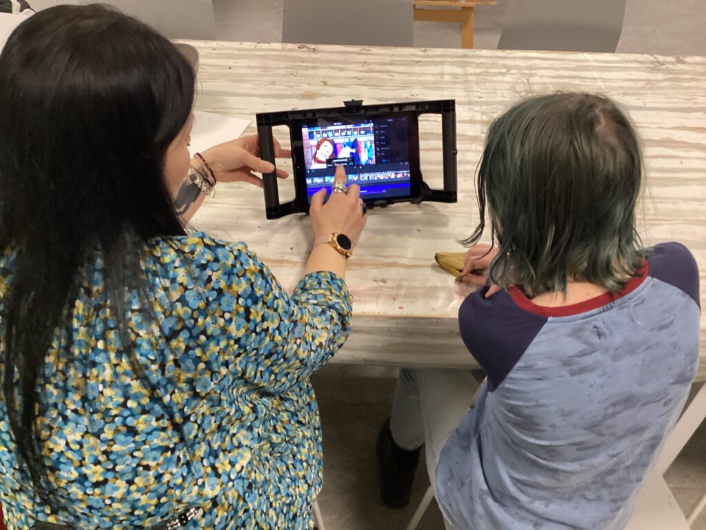 En vuxen med långt brunt hår och blommig tröja visar något på en skärm  för ett barn med långt hår och blå tröja. Båda sitter med ryggen mot.