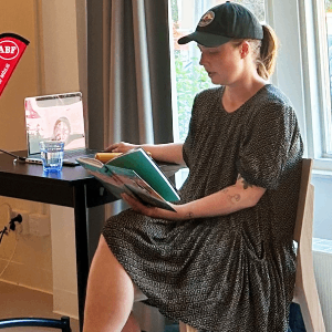 En person sitter och läser ur en bok vid ett bord framför en stor skärm som visar en illustration av en stol. I bakgrunden syns en röd ABF-banderoll.