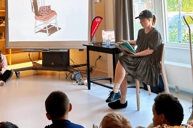 En person sitter och läser ur en bok vid ett bord framför en stor skärm som visar en illustration av en stol. I bakgrunden syns en röd ABF-banderoll.