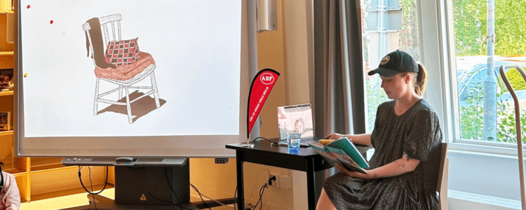 En person sitter och läser ur en bok vid ett bord framför en stor skärm som visar en illustration av en stol. I bakgrunden syns en röd ABF-banderoll.