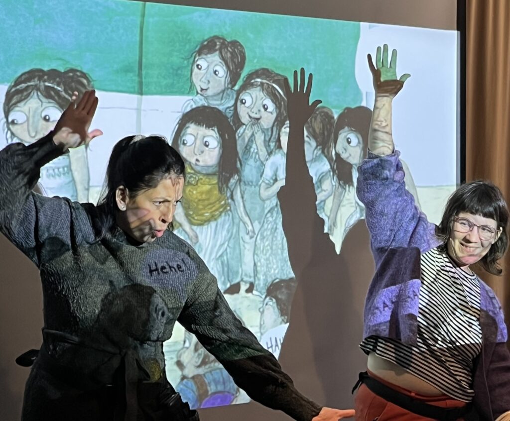 Två kvinnor dansar framför projicerad bild av barn som står och tittar på något.