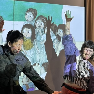 Två kvinnor dansar med stora rörelser framför en projektorvisad bild på barn från en sagobok.