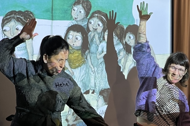 Två kvinnor dansar med stora rörelser framför en projektorvisad bild på barn från en sagobok.