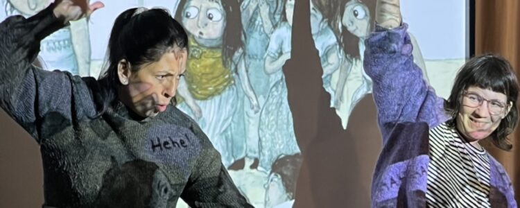 Två kvinnor dansar med stora rörelser framför en projektorvisad bild på barn från en sagobok.