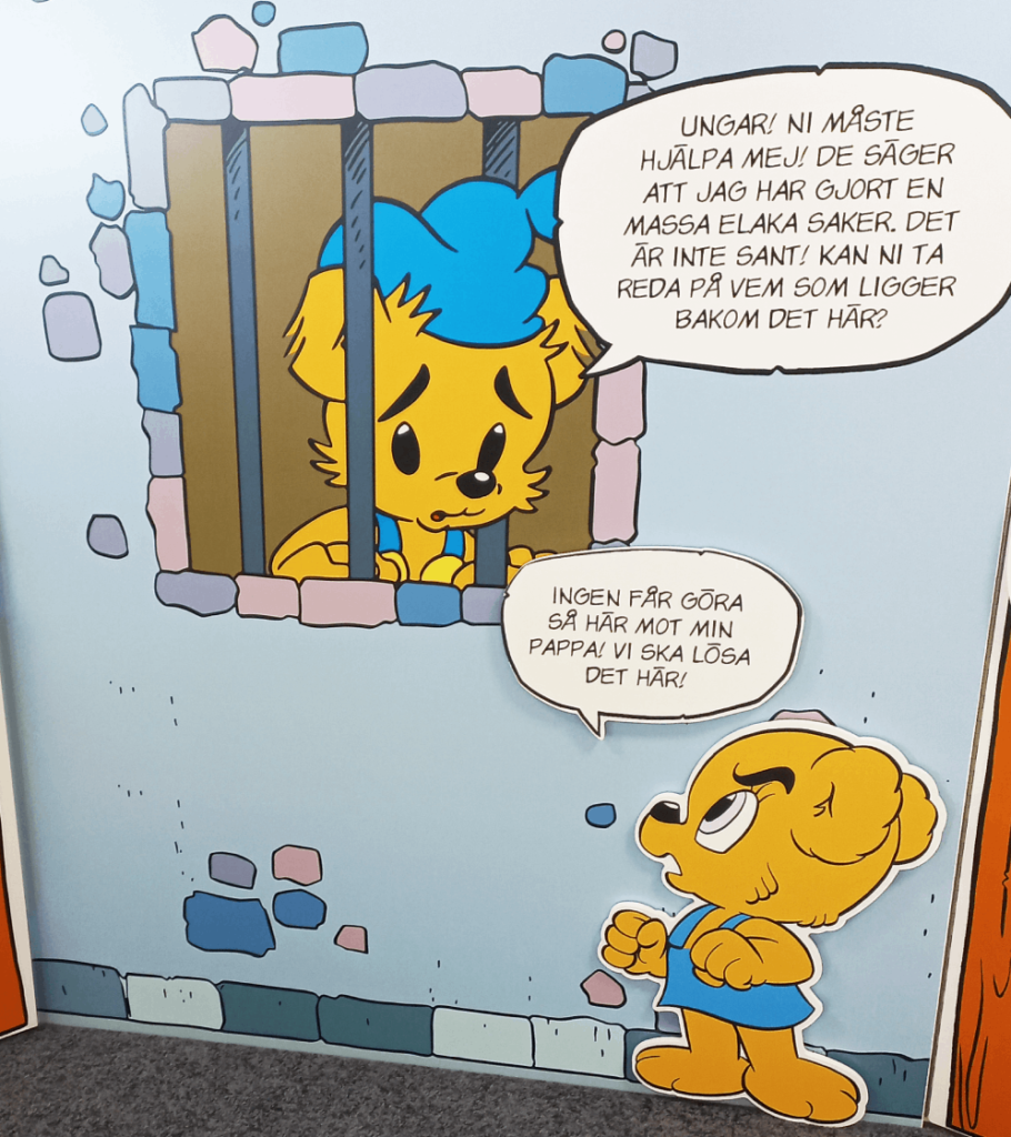 Närbild på en panel från utställningen "Bamses Källspanare". Bamse sitter bakom galler och säger: "Ungar! Ni måste hjälpa mej! De säger att jag har gjort en massa elaka saker. Det är inte sant! Kan ni ta reda på vem som ligger bakom det här?" Nedanför står Nalle-Maja
och svarar: "Ingen får göra så här mot min pappa! Vi ska lösa det här!"
