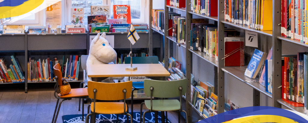 Barnavdelning på ett bibliotek med bokhyllor, små stolar och ett bord där en Muminfigur sitter bredvid en liten sverigefinsk flagga.