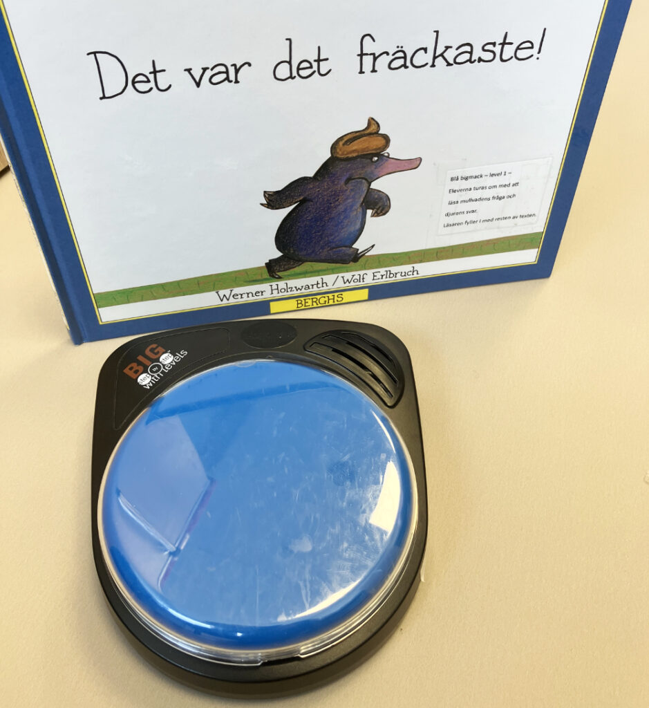 
Boken "Det var det fräckaste" av Werner Holzwarth och Wolf Erlbruch, med en illustration av en mullvad på omslaget, står uppställd på ett bord. Framför boken ligger en stor blå kommunikationsknapp (Big Step-by-Step med levels).