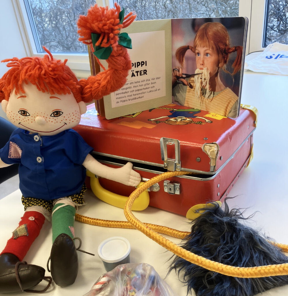 En Pippi Långstrump-docka med röda flätor och blå jacka sitter framför en röd resväska. På väskan står en uppslagen bok med texten "Pippi äter" och ett fotografi av Pippi som äter pasta. 
