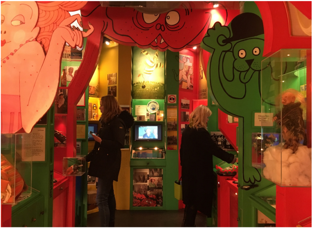 Färgglad museiutställning med illustrerade seriefigurer på väggarna. Två besökare tittar på montrar och en skärm som visar film.