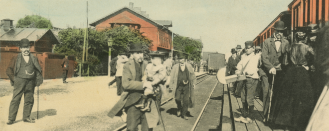 Handkolorerat historiskt foto från järnvägsstationen i Svedala, med män i sekelskifteskläder på perrongen.