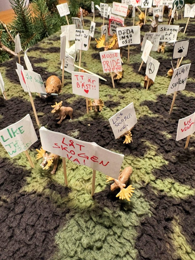 Miniatyrdiorama av en skog där små älgfigurer demonstrerar med handgjorda skyltar på tandpetare, bland annat "Låt skogen va!", "Moose pride" och "Döda inte oss!".