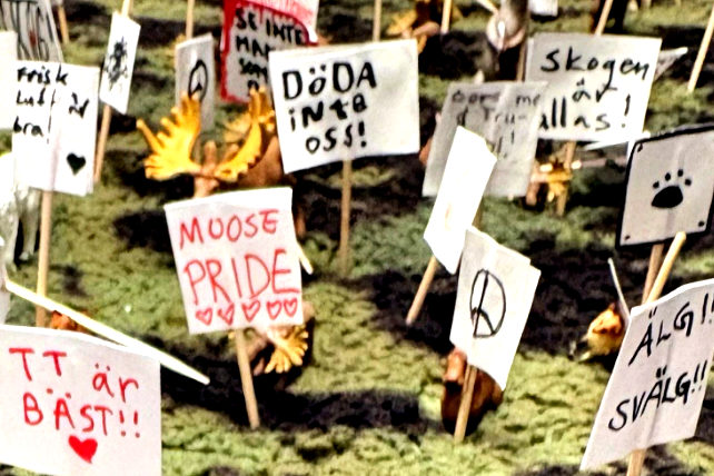 Miniatyrdiorama av en skog där små älgfigurer demonstrerar med handgjorda skyltar på tandpetare, bland annat "Låt skogen va!", "Moose pride" och "Döda inte oss!".